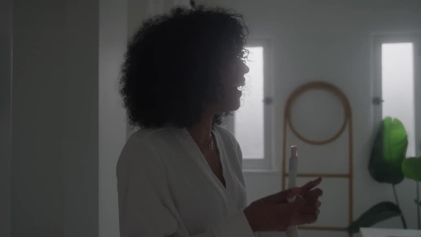 A woman with dark voluminous curly hair wearing a white robe and a delicate necklace stands in a brightly lit room near a window and a round mirror holding a bottle and laughing