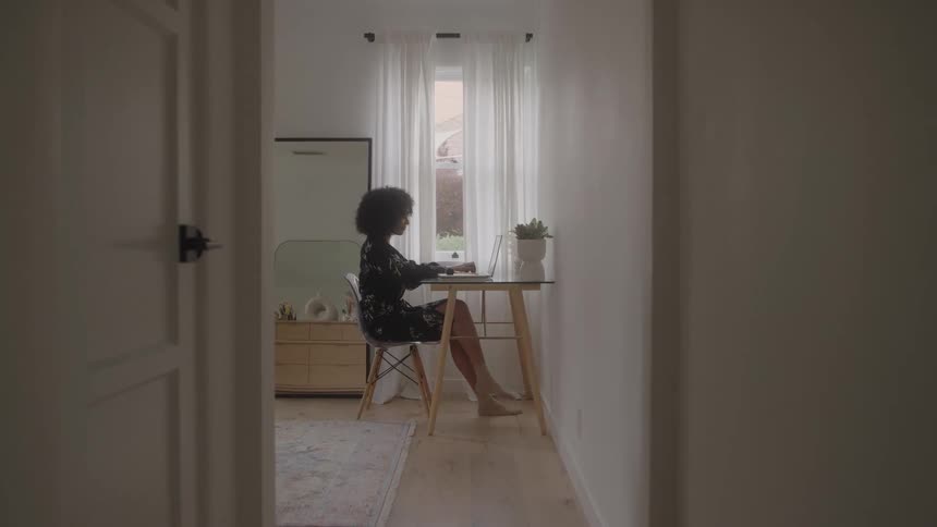 Viewed from a hallway a woman sits in profile working on her laptop at a small desk pausing to brush back her hair in the bright light of a window