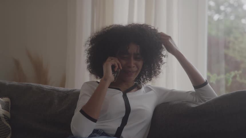 A woman with curly hair relaxes on her couch while smiling and laughing during a pleasant phone conversation her hand brushing through her hair