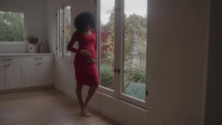 Pregnant woman in red dress looking out the window in softly lit indoor setting