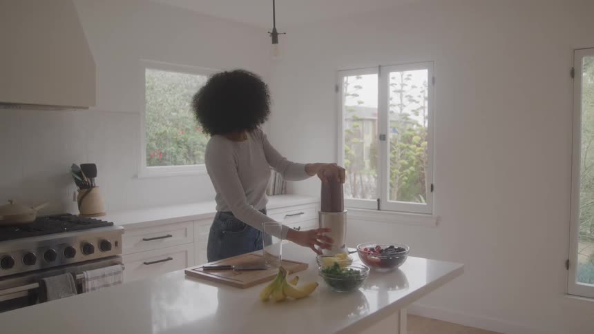 A woman is standing at a clean white kitchen island operating a blender to make a smoothie bowls of various fruits and greens including bananas and berries are laid out on the counter suggesting healthy meal preparation