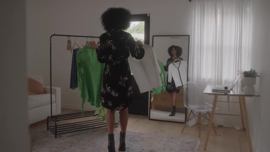 A woman in a black floral dress walks over to a clothing rack in a bright room browses the items and pulls out a white garment to examine in the mirror