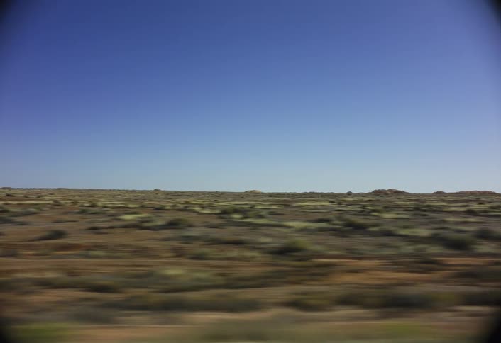 The camera captures a sweeping view of a vast flat desert or tundra landscape under a clear blue sky the frame is dominated by the dry lowlying ground which is a mix of brown yellow and green scrub moving quickly past the viewer suggesting travel