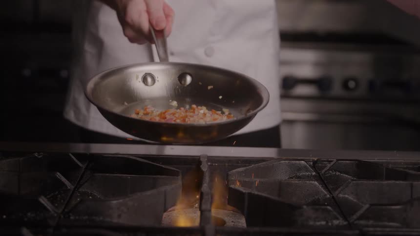 A chef wearing a white uniform is captured midmotion holding a silver saut pan filled with diced vegetables just above a lit commercial gas burner