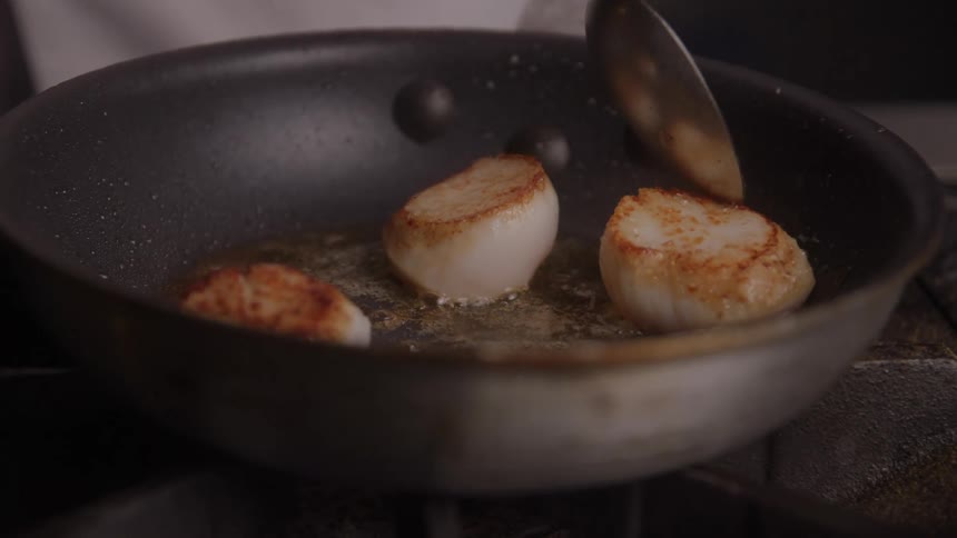 A persons fingers and a spoon are visible above a dark nonstick pan turning three large goldenbrowned sea scallops sizzling in butter