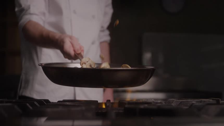 Chef standing next to a stove flipping sliced mushrooms in a frying pan over the flames