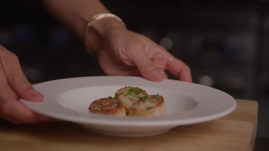Hands picking up a plate with seared scallops garnished with herbs