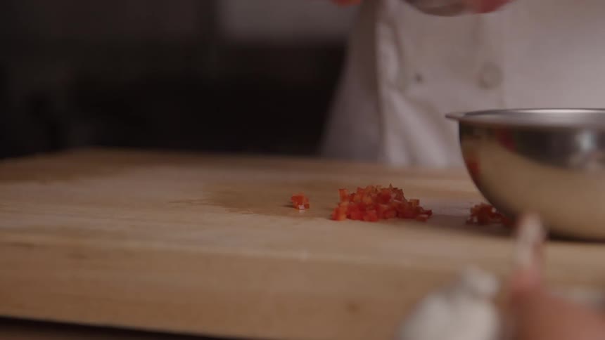 A persons hands use a small knife mincing to scoop diced orange vegetables into a metal bowl