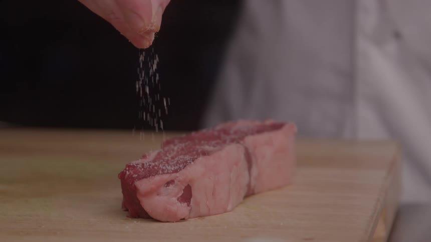 Chef sprinkles spices on a piece of raw meat on the kitchen counter