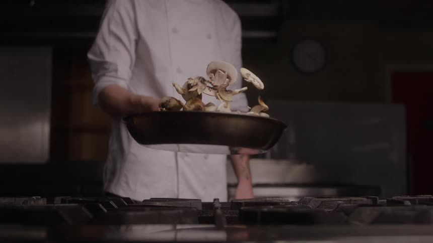Cook standing over a stove flipping sliced mushrooms in a frying pan