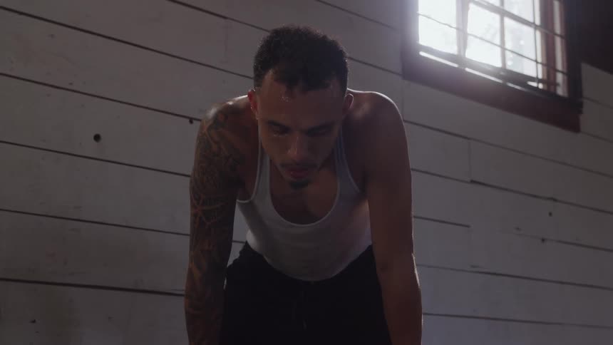 A young hispanic man wearing a singlet stands indoors at midday with a sweaty face and tattooed body looking exhausted as he catches his breath
