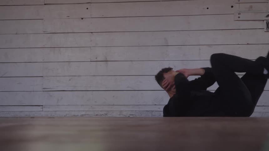 A man with curly hair dressed entirely in black is performing an abdominal crunch exercise on a wooden floor against a textured wall made of distressed white horizontal wooden planks
