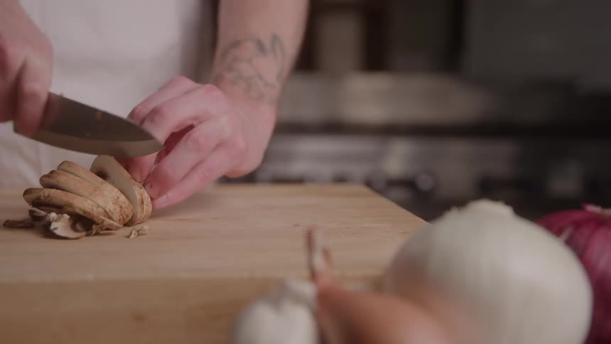 A person with a prominent forearm tattoo and a white apron is concentrating on slicing brown mushrooms with a large chefs knife on a wooden cutting board with onions and garlic cloves resting in the blurred foreground