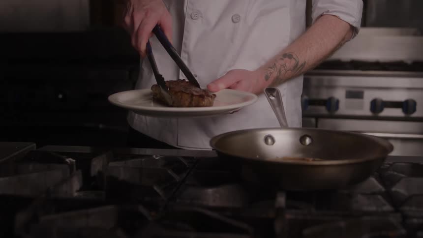 Cook holds his tongs as he takes piece of meat out of the frying pan and puts it on a plate