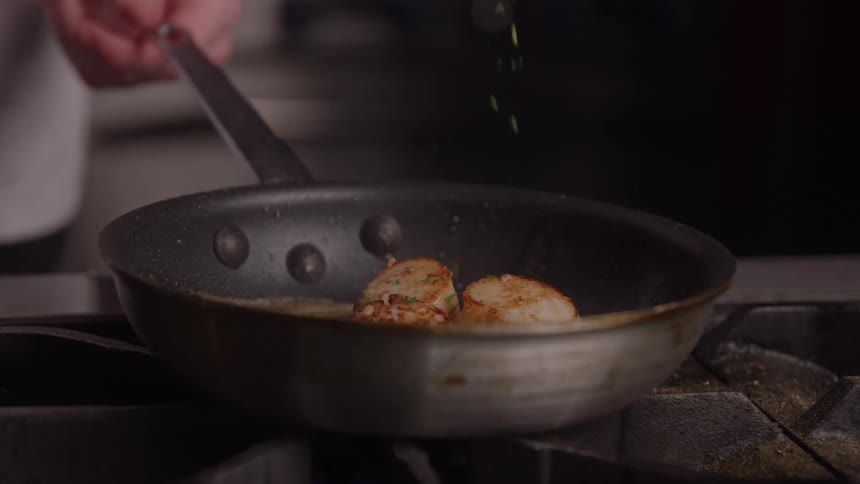 Herbs being sprinkled over seared scallops in the pan