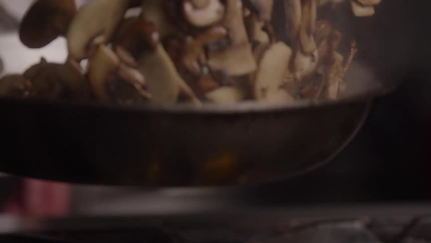 A person is vigorously tossing a generous quantity of sliced mushrooms in a stainless steel frying pan causing the contents to rise and fall while cooking over a visible gas flame creating steam and smoke