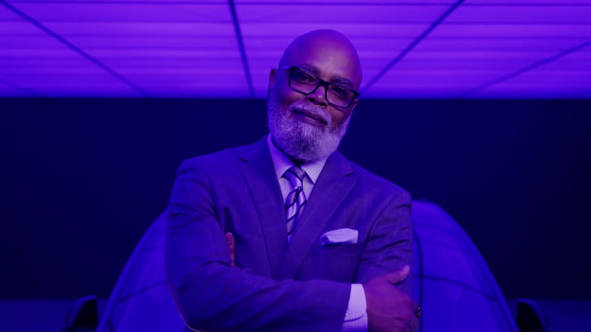 A confident well dressed man with a grey beard stands with his arms crossed in a futuristic room with dramatic purple lighting