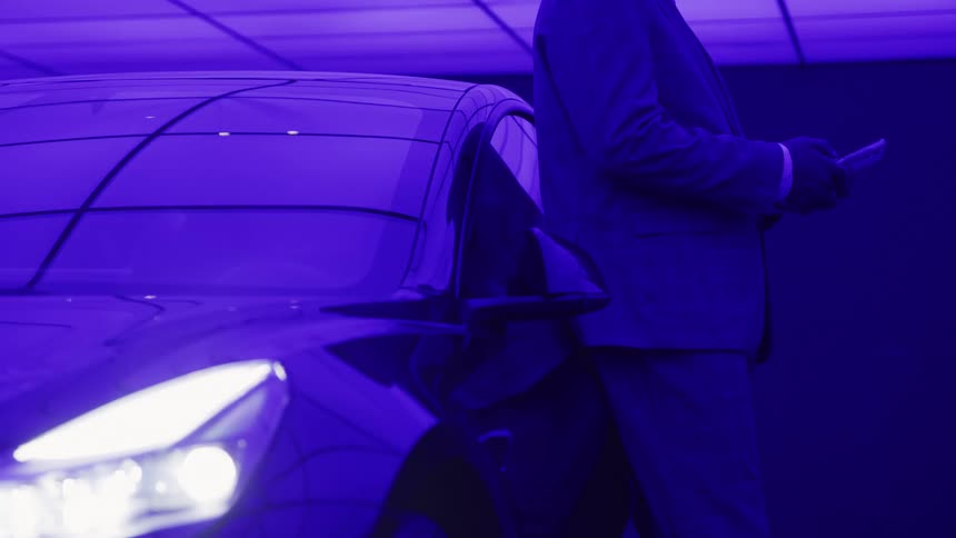 A person dressed in a dark suit and gloves is standing motionless next to the reflective illuminated front fender of a sleek modern car all bathed in an intense monochromatic purpleblue light