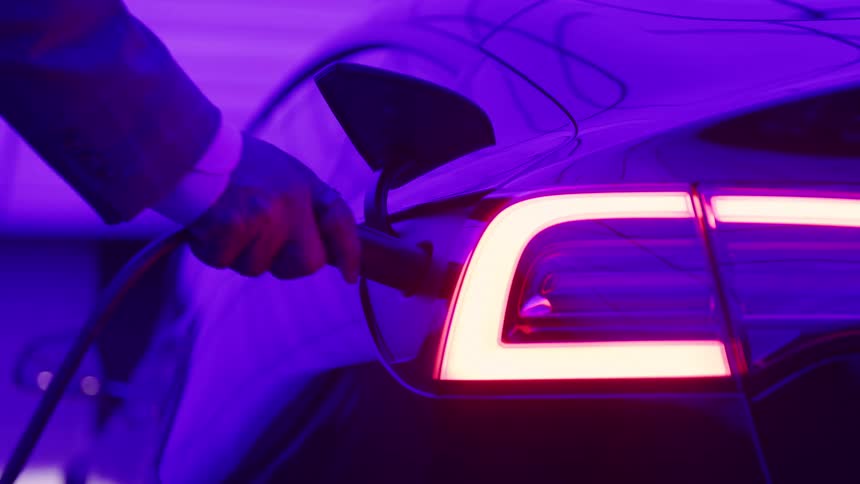 A hand wearing a suit jacket and white shirt cuff is seen plugging a black charging cable into the charging port of a sleek electric car bathed in dramatic neon purple and magenta light that highlights the bright angular rear taillight