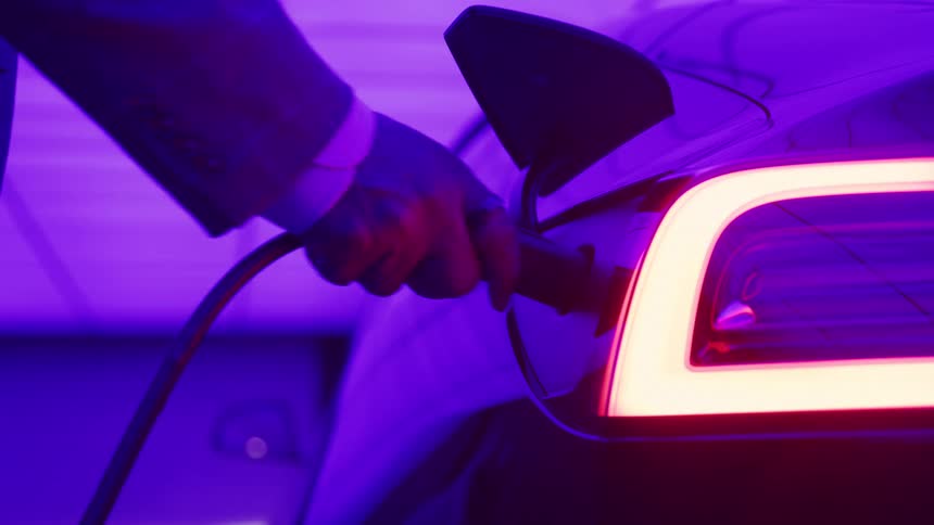 A hand in a suit jacket plugs a charging cable into an electric cars port bathed in purple light as the taillight glows brightly