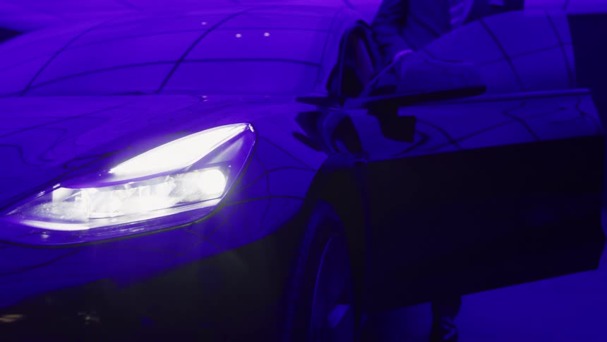 Under the dramatic intense glow of a deep purple ambient light the gleaming dark surface of a modern car is highlighted with its bright white headlight focused forward as an unseen driver appears to be opening the door and sitting in the drivers seat