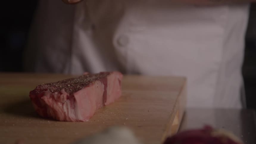 Chef sprinkles spices on a piece of raw meat on the kitchen counter