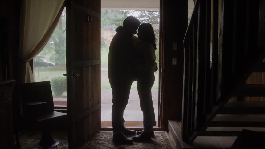 A couple is standing in a heartfelt embrace in a doorway their figures sharply silhouetted against the bright light of the exterior contrasting with the dark wooden tones of the interior staircase and furniture that frame them