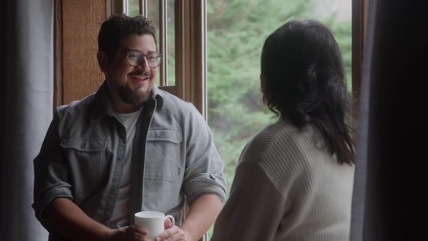 A man and a woman indoors next to a window are talking as the man holds onto a white mug