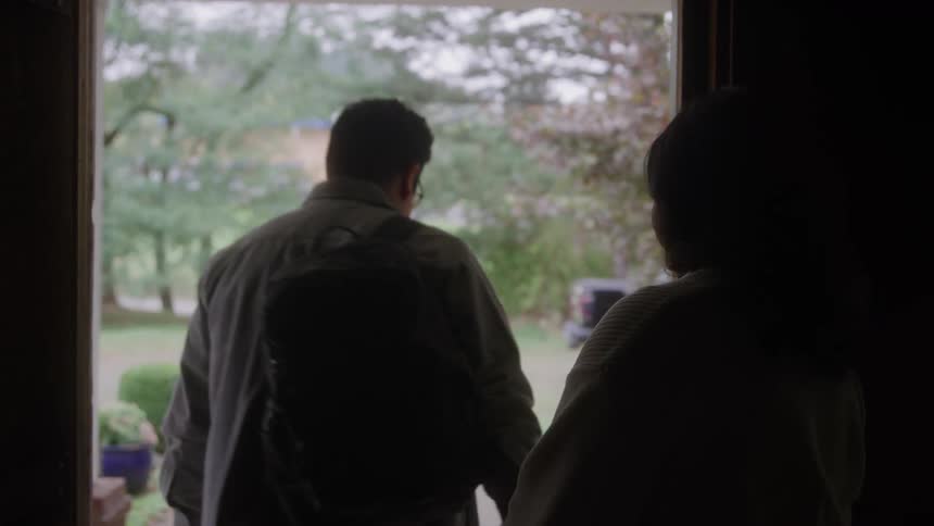 A man and a woman are framed in a dark doorway sharing a moment before the man wearing a backpack turns to walk away from the threshold into a bright green outdoor setting while the woman remains watching from the interior