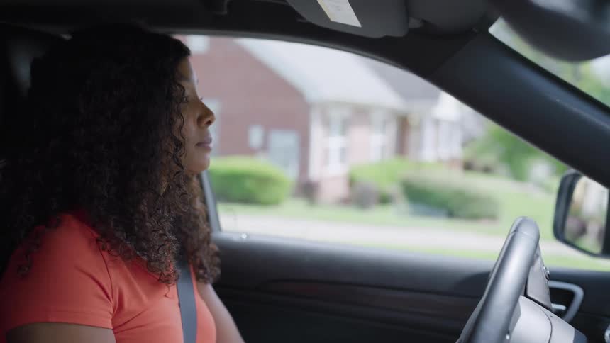 A woman in an orange shirt drives a car through a suburban neighborhood looking over her shoulder briefly before turning her attention back to the road