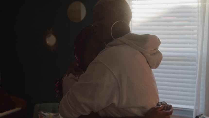 Adult black woman wearing a hoodie hugging a black teenager girl wearing glasses in a room indoors during day time