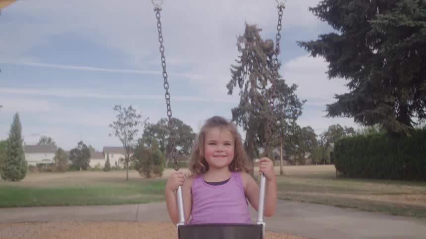 A young girl with a happy expression is swinging on a bucket seat swing on a sunny day moving toward and away from the camera in a public park with grass trees and distant houses visible in the background