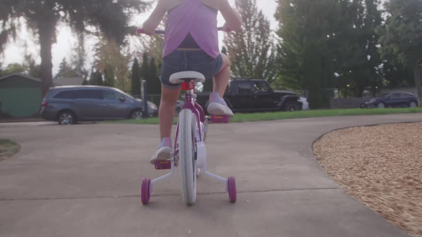 A young girl pedals her bike with training wheels along a paved path in a park