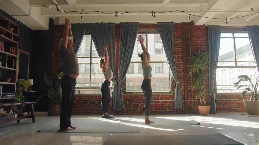 Two young women and a middleaged woman practice yoga stretches together indoors at midday each on their own mat
