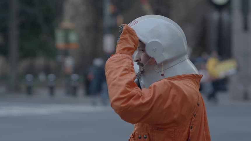 A man wearing an orange jumpsuit and a helmet standing outside by a city street and looking around