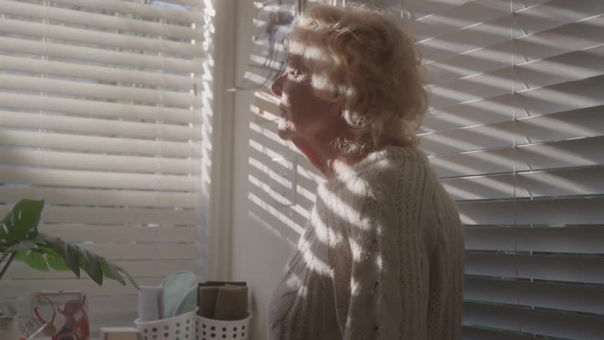 Woman with her eyes closed receives light filtered through the blinds inside her home