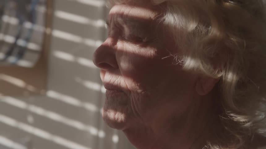 Elderly woman with closed eyes receiving light filtered through window blinds