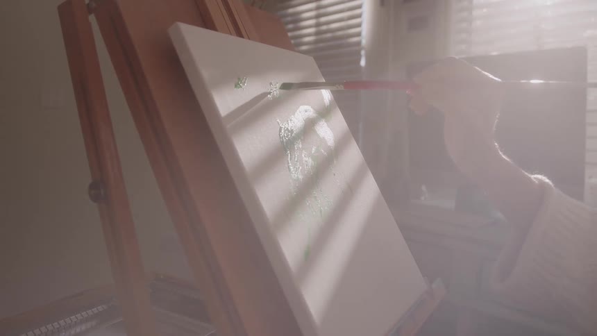 An artist delicately applies green paint to a canvas on an easel in a room filled with hazy atmospheric light
