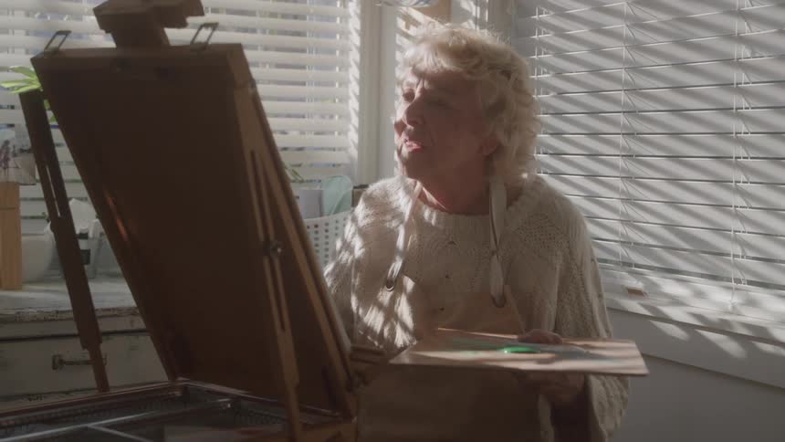 Elderly woman painting at her easel under light filtered through the blinds