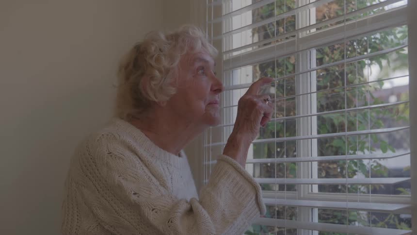 An elderly woman with grey hair dressed in knitwear opens the window blind with her fingers at midday gazing outside at a tree