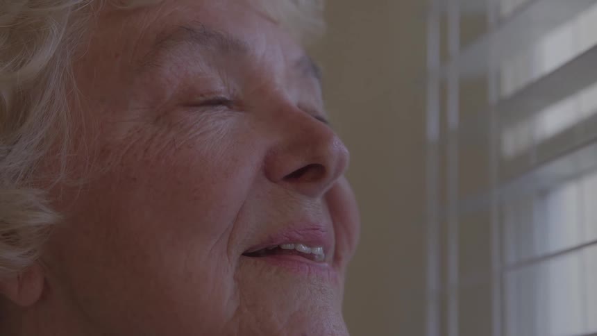 Elderly woman in profile looking outside through white blinds with natural light