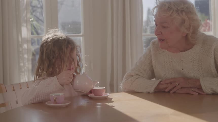 Girl pours tea for an older woman using a pink porcelain set lit by sunlight