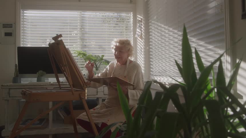 An elderly woman with short curly blonde hair wearing an apron is seen painting thoughtfully on an easel near a sunny window with blinds creating an artistic and serene mood indoors