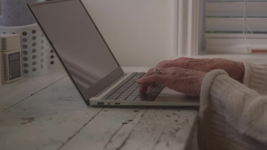 A closeup shot captures the wrinkled hands of an older person who is actively typing on the keyboard of a modern silver laptop resting on a rustic white wooden desk near a window