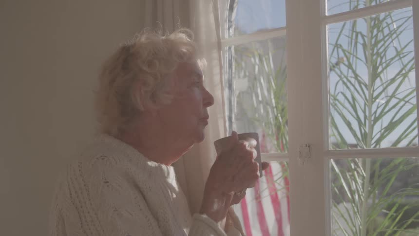 An elderly woman with curly white hair peacefully sips a warm drink while gazing out a sunlit window