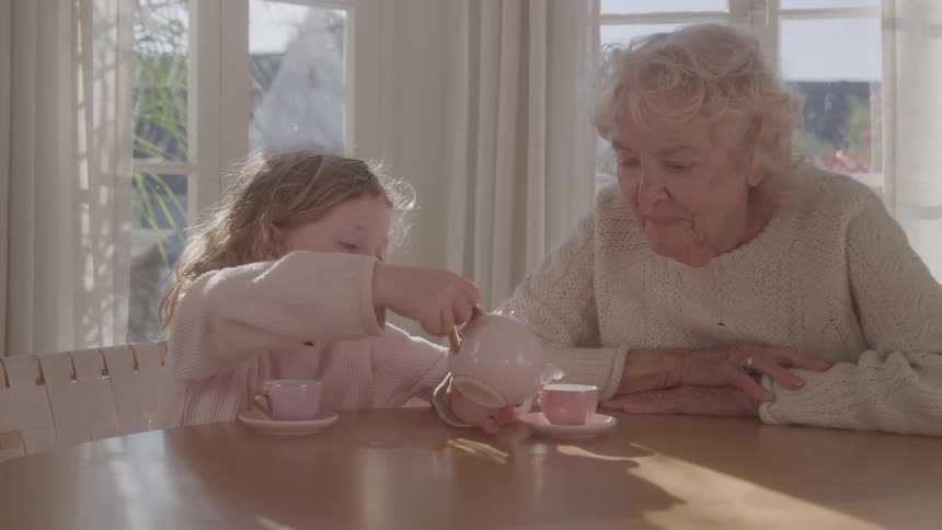 A grandmother sits beside her granddaughter at the dining table at midday watching as the little girl carefully pours tea from a teapot into a teacup