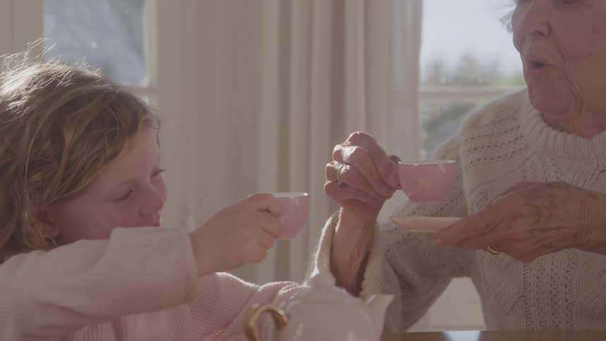 An elderly woman in knitwear wearing a gold wedding ring sits at the dining table at midday with a young girl in a sweatshirt they smile warmly as they raise their teacups together in a cheerful toast