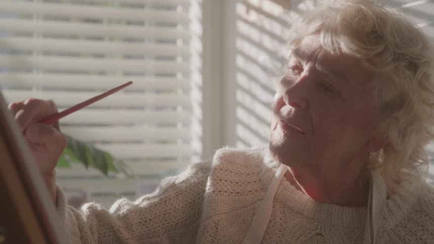 Elderly woman painting a canvas lit by window light