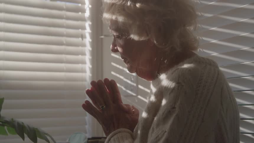 Elderly woman with hands together in a contemplative gesture near the window