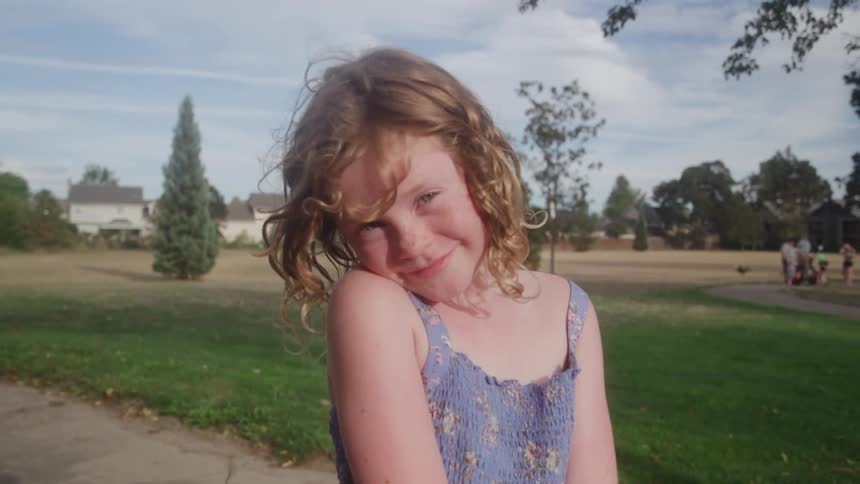 A shy little girl with curly red hair smiles sweetly while standing in a park on a sunny day